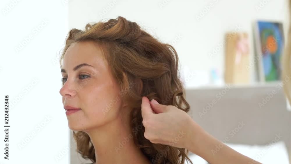 Hairdresser using hairspray while creating curly hairstyle to long haired woman. Hairstylist making curling hair to fashion model. Woman face during hairstyling. Hairdresser using hairspray while creating curly hairstyle to long haired woman. Hairstylist making curling hair to fashion model. Woman face during hairstyling.