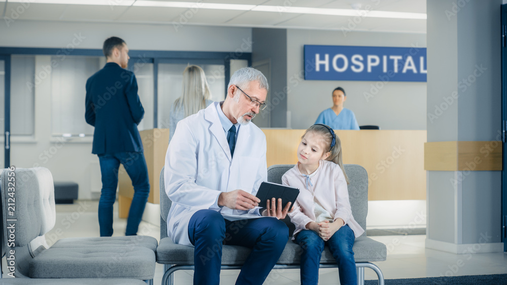 In the Lobby of the Hospital Male Doctor Talks with a Cute Little Girl while Sitting on the Sofa, He's Showing Her Tablet Computer. Busy Modern Hospital. In the Lobby of the Hospital Male Doctor Talks with a Cute Little Girl while Sitting on the Sofa, He's Showing Her Tablet Computer. Busy Modern Hospital.