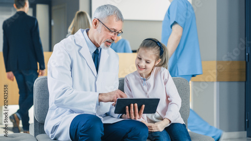 In the Lobby of the Hospital Male Doctor Talks with a Cute Little Girl while Sitting on the Sofa, He's Showing Tablet Computer to Her. Busy Modern Hospital with Best Pediatrics Department. 