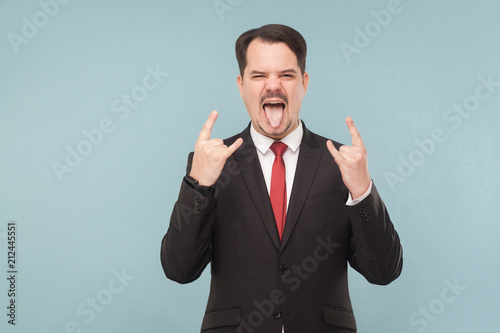 Emotional businessman showing rock sign. indoor studio shot. isolated on light blue background. handsome businessman with black suit, red tie and mustache looking at camera.