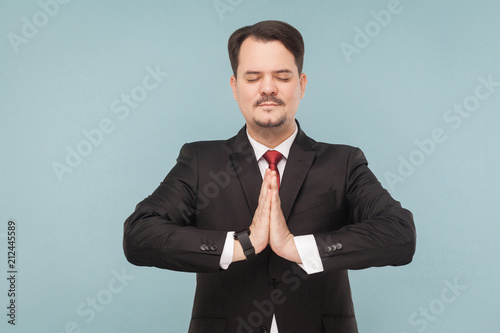 During a work break, it is best to do yoga to put your nerves in order. indoor studio shot. isolated on light blue background. handsome businessman with black suit, red tie and mustache.