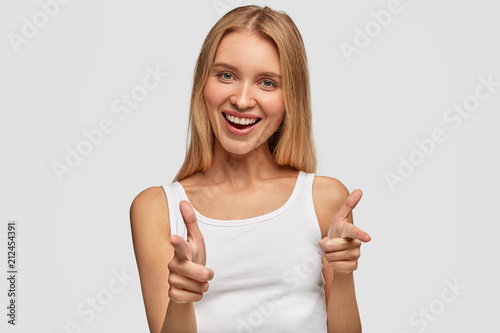 Obraz na plátne Portrait of happy cute blonde female with joyful expression, points at you with