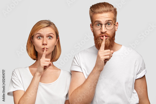 Horizontal shot of two girlfriend and boyfriend make silence gesture, ask not tell about their secret and be quiet, have surprsied expressions, isolated over white wall, touch lips with index fingers
