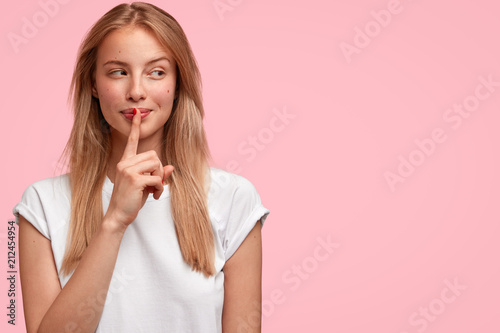 Indoor shot of beautiful female looks mysteriously aside, has intriguing look, asks to be quiet, dressed in casual clothes, stands against pink background with blank copy space for your advertisement photo