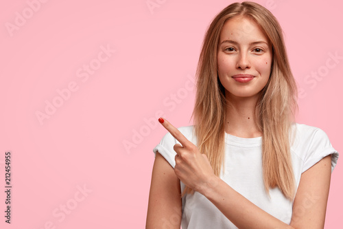 Studio shot of beautiful blonde female with satisfied expression, points with fore finger aside, shows copy space for your advertisement or promotional text, has appealing look and pure skin