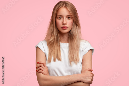 Confident attractive young European female keeps hands crossed, looks seriously directly at camera, contemplates about future plans after graduation university, isolated over pink background