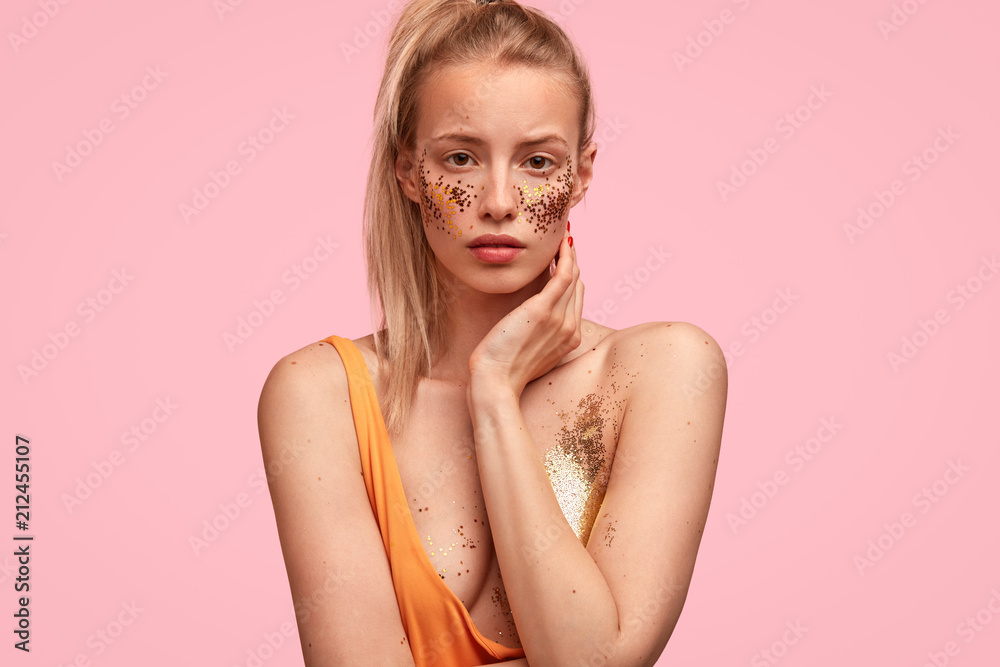 Lovely self confident female has festive make up, looks seriously, prepares for party with friends, has bare shoulders, thoughtful expression, isolated over pink background. Woman with glitter on skin
