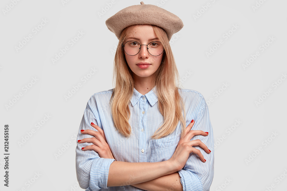 Horizontal shot of pretty lovely young blonde female keeps arms folded, dressed in elegant shirt and beret, has retro style in clothes, has clever expression, stands against white background. Horizontal shot of pretty lovely young blonde female keeps arms folded, dressed in elegant shirt and beret, has retro style in clothes, has clever expression, stands against white background.