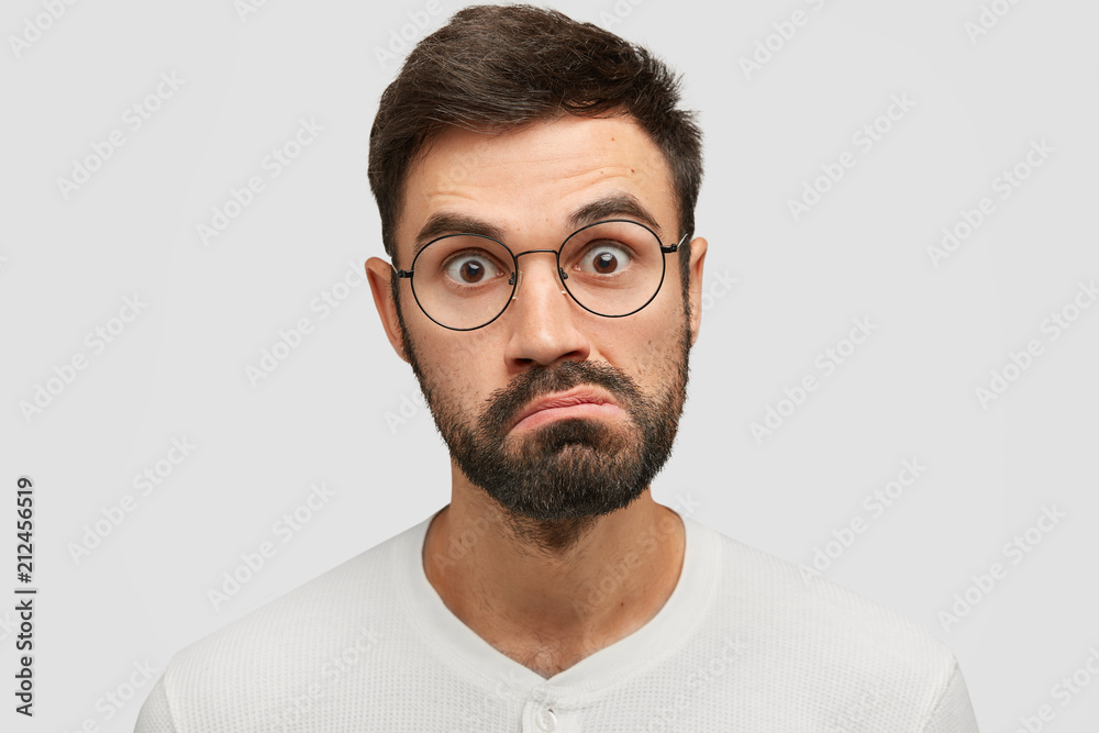 Obraz premium Close up portrait of handsome angry surprised Caucasian male looks in bewilderment, purses lips and stares at camera, wears glasses, isolated over white background. People, facial expressions