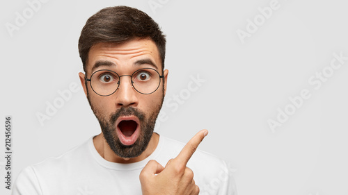 Close up shot of emotive shocked bearded young male opens mouth widely, stare at camera, points upwards aside at blank space, says: Wow, it`s something unbelievable! People and emotions concept