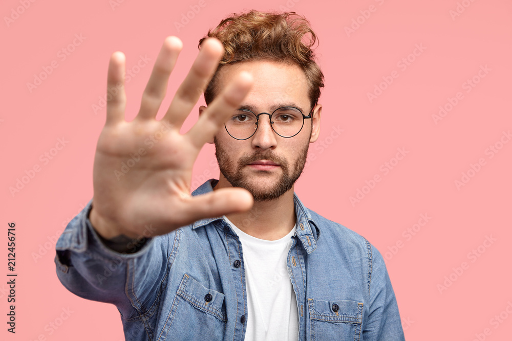 Serious handsome bearded hipster stretches hand towards camera, shows palm, demonstrates stop or hold gesture, wears fashionable clothes, stands against pink background. Stay away from me, please Serious handsome bearded hipster stretches hand towards camera, shows palm, demonstrates stop or hold gesture, wears fashionable clothes, stands against pink background. Stay away from me, please