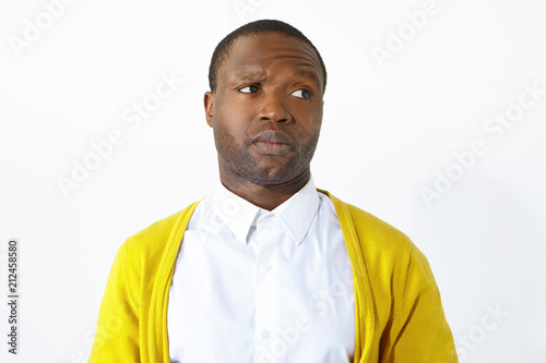Picture of irritated unhappy young dark skinned man frowning and looking sideways with grumpy expression, annoyed with disturbing sound, posing isolated against white studio wall background