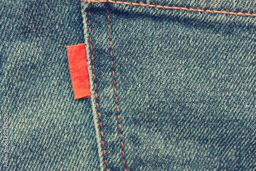 Vintage Style Image of Tag on Denim Jeans Background. Empty Red Label on Casual Pair of Jeans, Close Up Top View. Simple Empty Tag on Jean Texture, Retro Style Filter Effect.