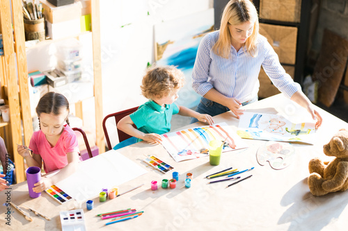 High angle portrait of young woman drawing with three little children sitting at wooden table in modern art studio for kids