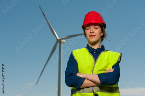Portrait of a handsome successful young woman the chief designer in a green waistcoat and a red helmet against the background of windmills and a blue sky.