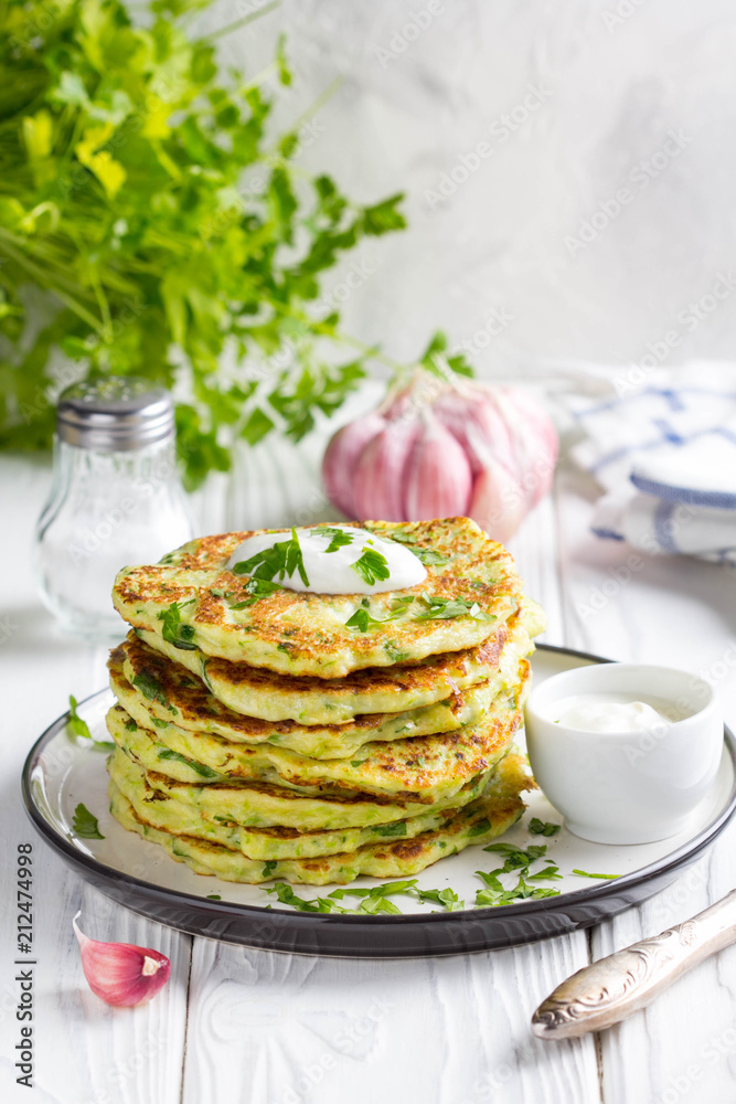Zucchini pancakes with parsley and sour cream, summer food, tasty snack. High stack in a plate on white wood Zucchini pancakes with parsley and sour cream, summer food, tasty snack. High stack in a plate on white wood