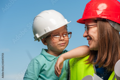 Cute little boy next to a positive mom with glasses and helmets on the background of blue skies. Concept of builders and architects