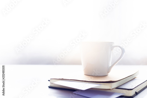 Coffee cup on notebook on white table in the morning sun light ,Working space