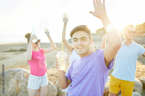 Excited guy with drink and his friends on background dancing on beach on summer day