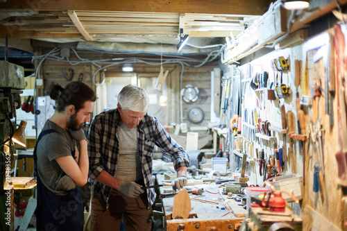 Woodworking masters standing by workbench while mature one processing wooden piece