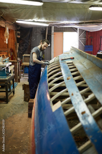 Young man in workwear standing by unfinished ship construction and working over it © pressmaster