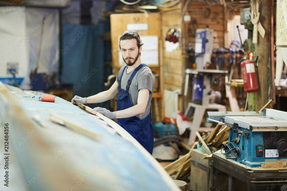 Serious young shipbuilder in workwear standing by large wooden vessel and repairing it Serious young shipbuilder in workwear standing by large wooden vessel and repairing it