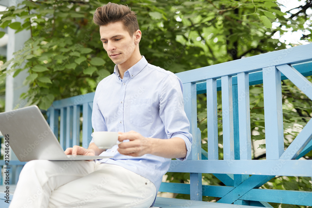 Fototapeta premium Young man with cup of coffee sitting on bench in summer cafe with laptop in front and searching for online ideas