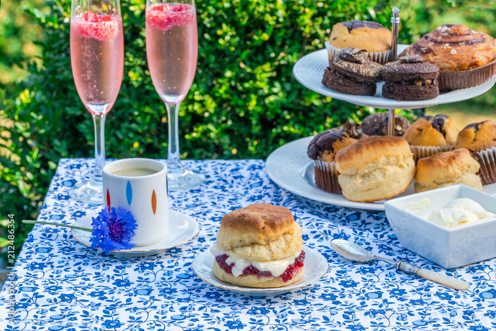 Afternoon tea with cakes and traditional English scones with strawberry jam and clotted cream set up on a table in the garden. Outdoor dining. Afternoon tea with cakes and traditional English scones with strawberry jam and clotted cream set up on a table in the garden. Outdoor dining.