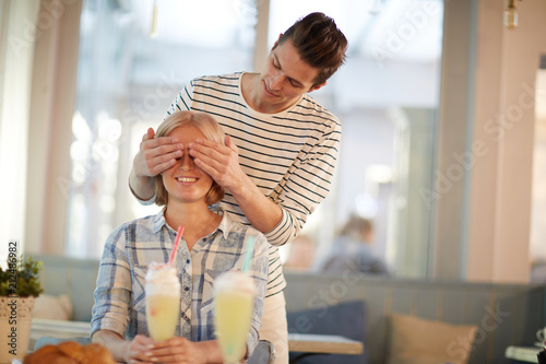 Young man covering his girlfriend eyes to make her surprise while sitting in cafe