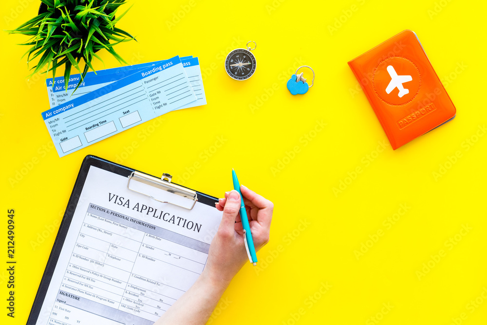 Hand fills visa application form. Form near glasses, pen, passport cover with airplane signand airplane tiskets on yellow background top view copy space Hand fills visa application form. Form near glasses, pen, passport cover with airplane signand airplane tiskets on yellow background top view copy space