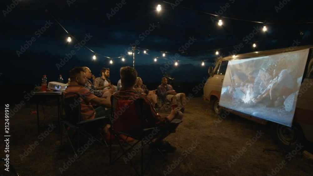 Friends gathering in campsite around bonfire and watching movie with projector on van side in dark evening