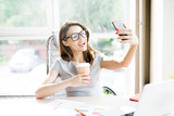 Portrait of beautiful businesswoman wearing glasses taking selfie  with coffee cup while working at desk in office  sitting against window in sunlight