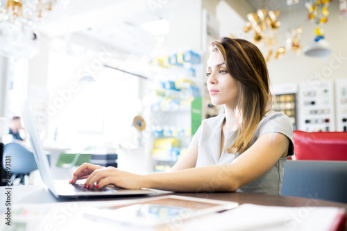 Side view portrait of beautiful young woman using laptop while sitting at desk in shop waiting for clients, copy space