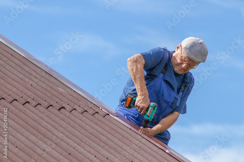 Old man working at heat on a roof of a house with electric screwdriver, wearing no safety devices, work clothing, blue overall, dangerous job, elder people at labor concept