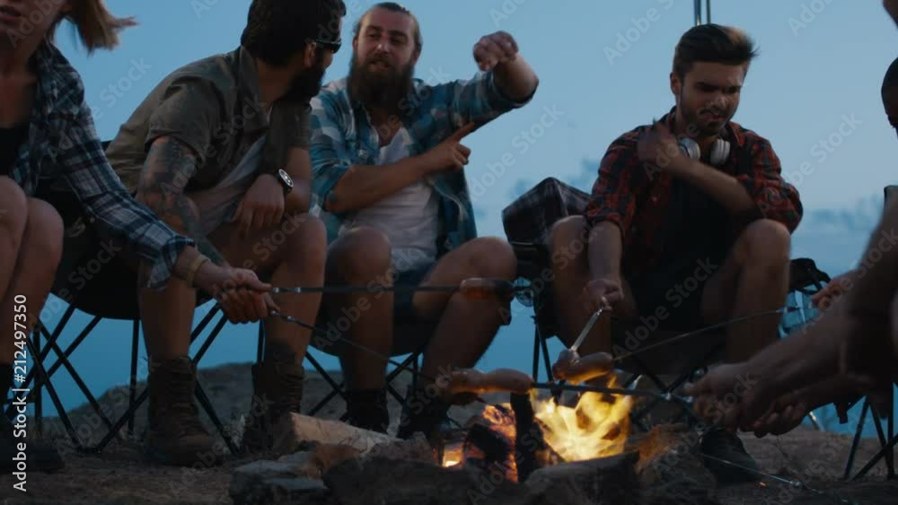 Group of cheerful friends relaxing in campsite and frying sausages on burning flame of bonfire gathering around and having fun Group of cheerful friends relaxing in campsite and frying sausages on burning flame of bonfire gathering around and having fun