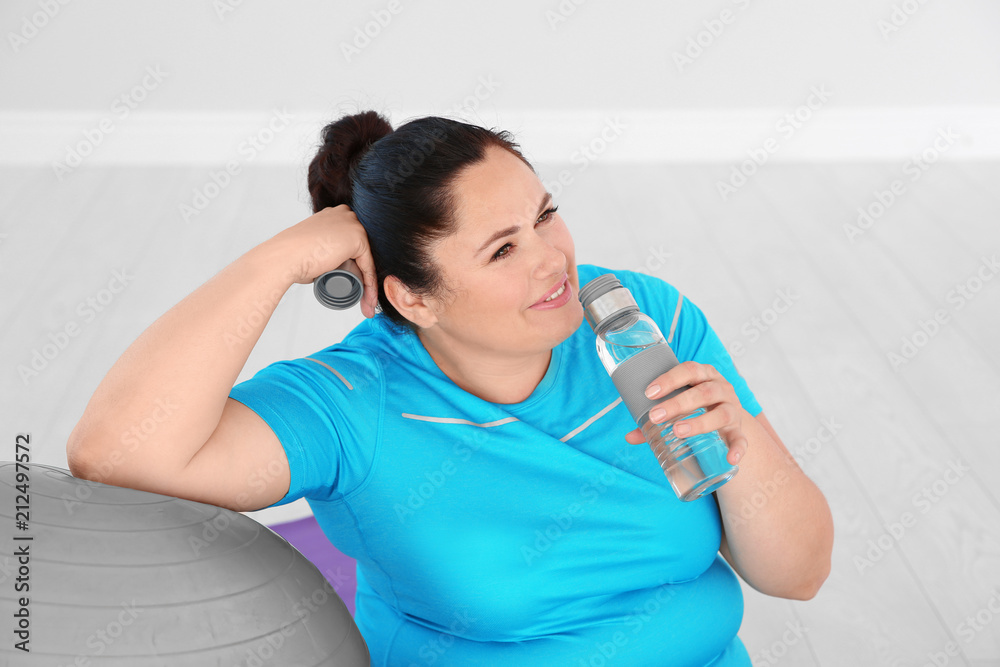 Tired overweight woman drinking water in gym