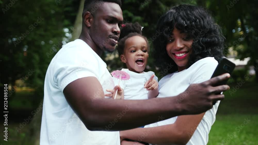 Cute african american family having fun in the park pose to take a selfie with mobile phone slow motion close up child girl daughter couple summer nature picnic mother parents portrait Cute african american family having fun in the park pose to take a selfie with mobile phone slow motion close up child girl daughter couple summer nature picnic mother parents portrait