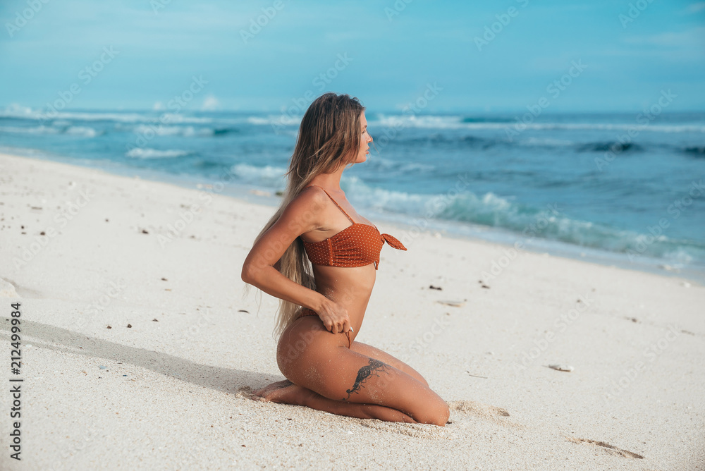 Side view, sexy model with a smooth skin and an even tan sitting on the lap on the beach against the blue sea, straightens his fashionable swimsuit, looks at the horizon. Side view, sexy model with a smooth skin and an even tan sitting on the lap on the beach against the blue sea, straightens his fashionable swimsuit, looks at the horizon.