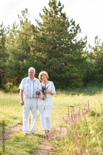 Elderly couple in the white linen dress standing outdoors, in the hands a bouquet of flowers, and on the background of coniferous forest © Alexandra