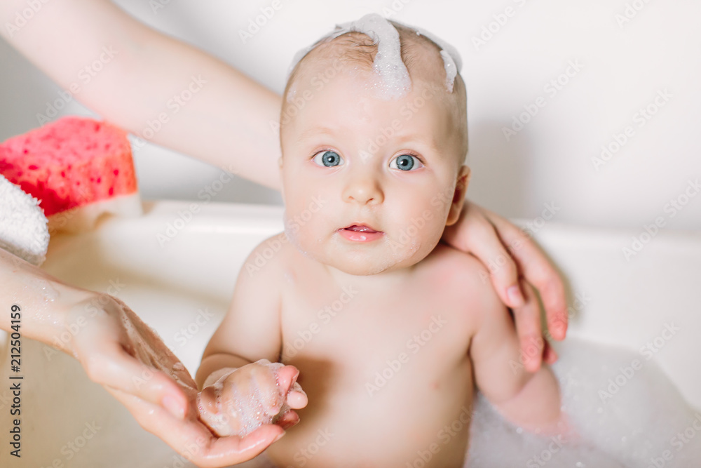 Happy laughing baby taking a bath playing with foam bubbles. Little child in a bathtub. Infant washing and bathing. Unrecognizable mother bathing her baby in white small plastic bat Happy laughing baby taking a bath playing with foam bubbles. Little child in a bathtub. Infant washing and bathing. Unrecognizable mother bathing her baby in white small plastic bat