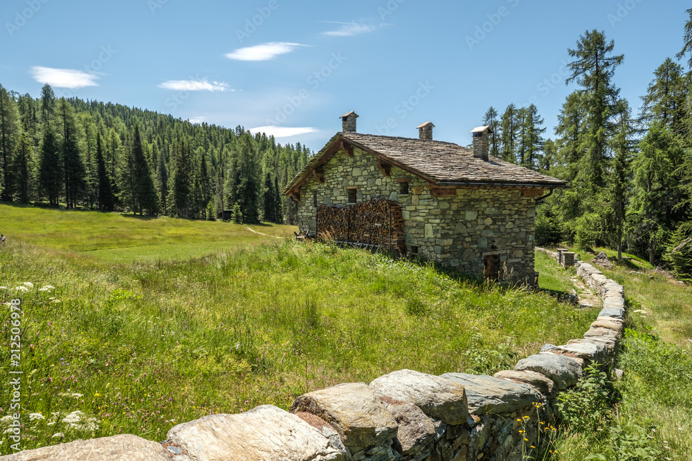 Rural mountain house with stone fence wall over a green yard against the forest and a blue sky. Mountain landscape in Valtellina, Italy. Rural mountain house with stone fence wall over a green yard against the forest and a blue sky. Mountain landscape in Valtellina, Italy.