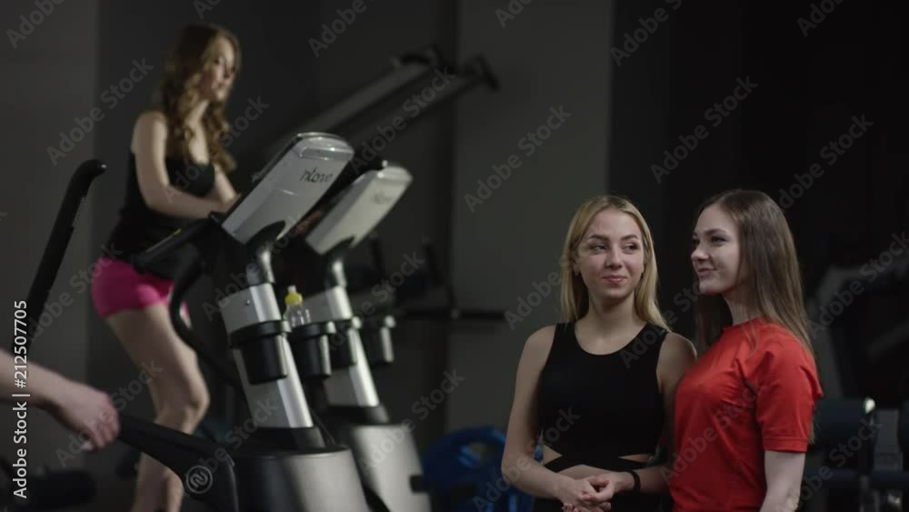 coach comes to greeting to pretty girls in new gym and high five and ask about them with girl engaged in simulators in the background coach comes to greeting to pretty girls in new gym and high five and ask about them with girl engaged in simulators in the background