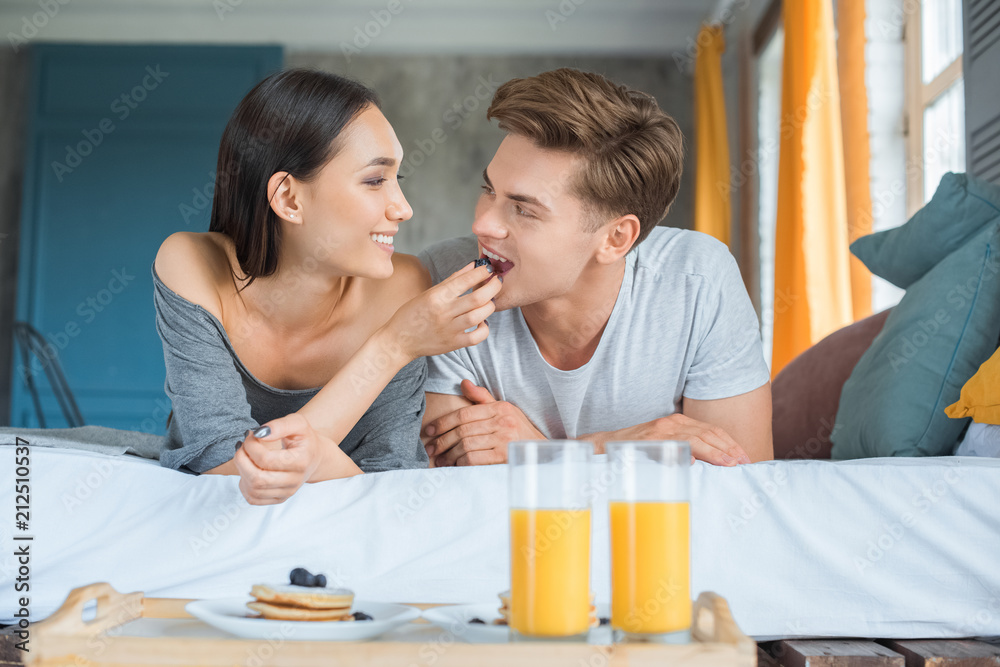 portrait of asian woman feeding caucasian boyfriend with breakfast in bed at home