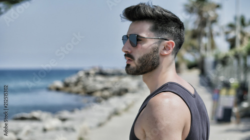 Attractive fit athletic young man soaking in the sun on seaside boardwalk or seafront, wearing black tank-top