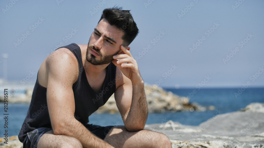 Attractive fit athletic young man soaking in the sun on seaside boardwalk or seafront, sitting on rock, wearing black tank-top
