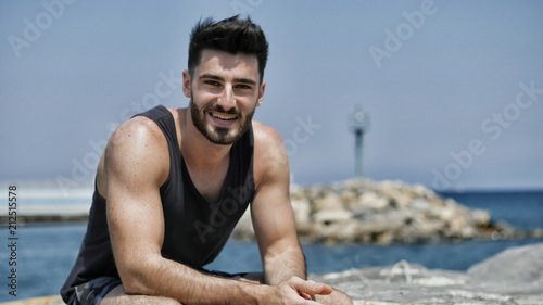 Attractive fit athletic young man soaking in the sun on seaside boardwalk or seafront, sitting on rock, wearing black tank-top, looking at camera with a smile