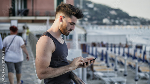 Fotografie Athletic young man at the seaside using cell phone to type message while looking