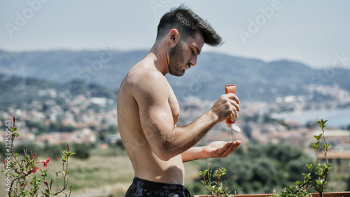 Shirtless Young Man Putting on Sunscreen Cream, Muscular Man Wearing Bathing Suit Ready to Sunbathe on Terrace