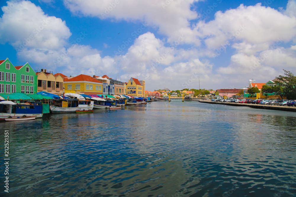 Obraz premium A view on the blue sea and many very colorful caribbean houses. Also there are some also colorful boats docking next to the houses. Furthermore there is a blue and white sky with some clouds.