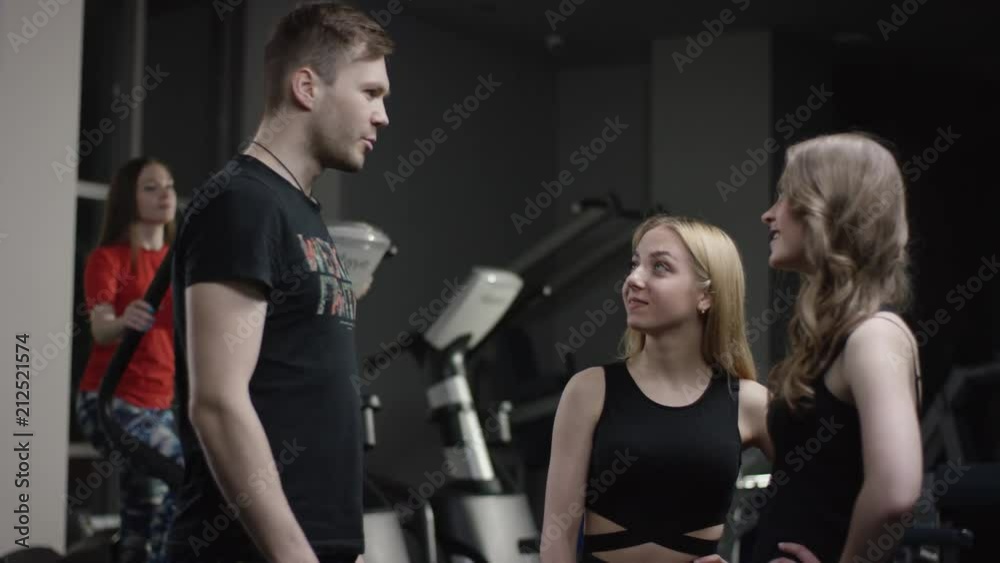 coach comes to greeting to beautiful girls in new gym and high five and ask about them with girl engaged in simulators in the background